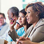 Mid adult African American businesswoman is answering a question during a job training seminar or business conference. She is wearing business casual clothing and holding a pen. Woman is sitting with diverse professional colleagues.