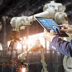 Man in a suit controls the arms of a welding robot from his tablet.