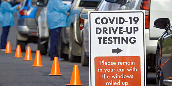 A large sign greets patients at a COVID-19 drive-up station as two female nurses in full protection gown and face mask attend to patients in their cars.