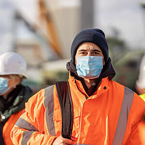 Head-on view of shipyard worker in a bright orange safety jacket and disposable mask, surrounded by out-of-focus co-workers.